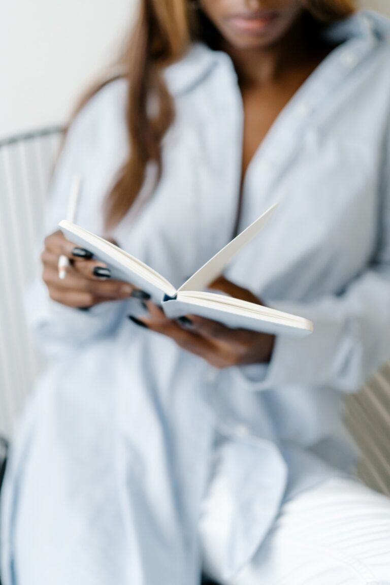 Woman sitting in a cozy chair wearing a white robe, holding an open journal—creating a peaceful moment to reflect on affirmations for each day of the week.