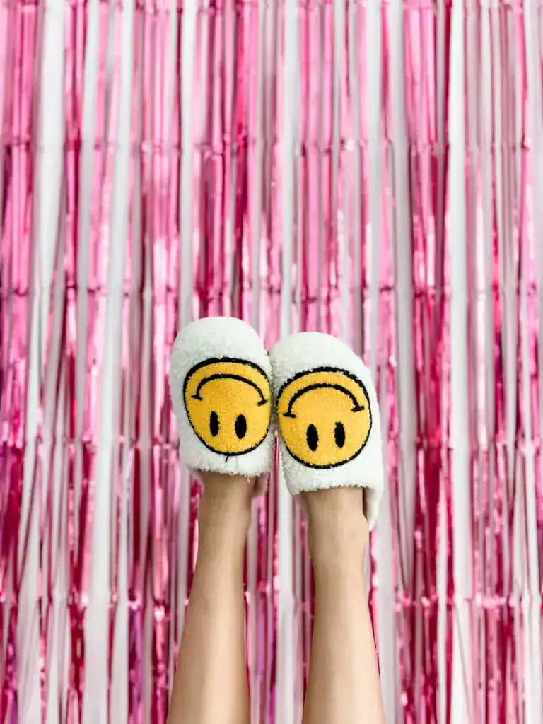Pink metallic fringe backdrop with feet wearing yellow smiley-face slippers, illustrating the playful freedom of things I no longer feel guilty about after 50.