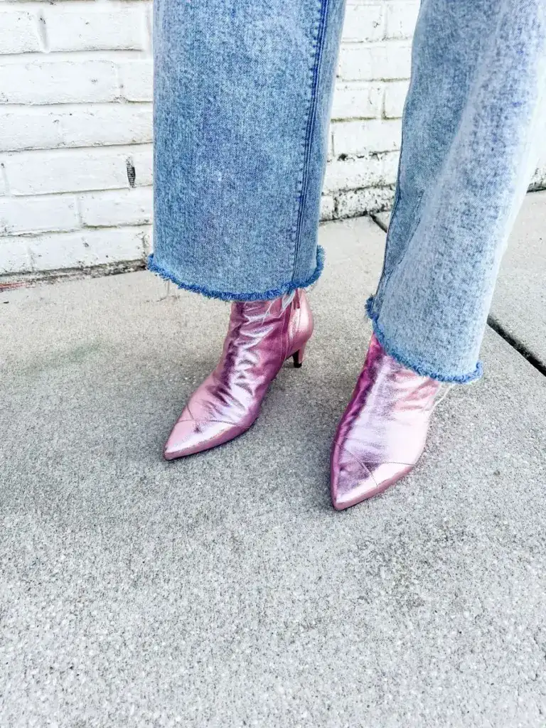 Metallic pink pointed-toe ankle boots styled with light-wash, frayed-hem jeans against a white brick wall.